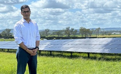  A man stands in a grassy field with rows of solar panels behind him. 